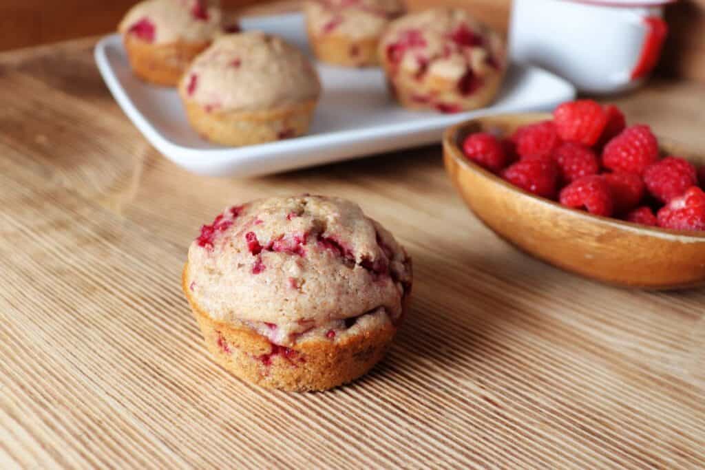 A raspberry spelt muffin sits on a board next to a wooden bowl full of fresh raspberries. A plate with more muffins is int he background. 