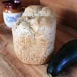 A loaf of bread sits on a board with a fresh zucchini next to it. In the background is a clay pitcher.