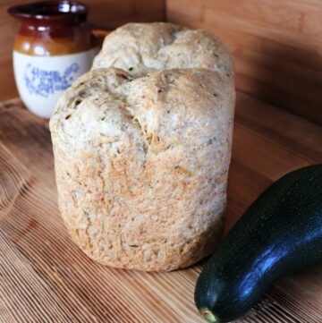 A loaf of bread sits on a board with a fresh zucchini next to it. In the background is a clay pitcher.