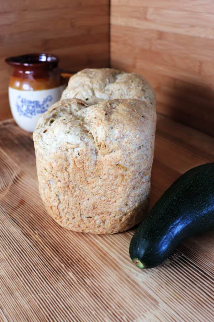 A loaf of bread sits on a board with a fresh zucchini next to it. In the background is a clay pitcher. 