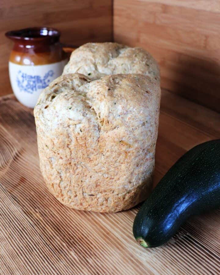A loaf of bread sits on a board with a fresh zucchini next to it. In the background is a clay pitcher.