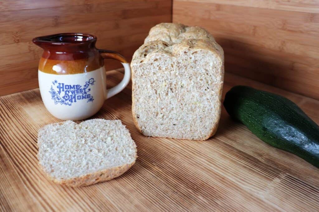 A loaf of bread with the end cut off sits on a board. The heel is sitting next to it. A fresh zucchini sits o the right of the bread. To the left is a clay pitcher that reads 'home sweet home'.