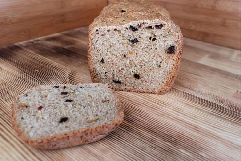 A loaf of zucchini raisin bread with the end cut off sits on a board. The heel of the bread sits in front of the loaf. 