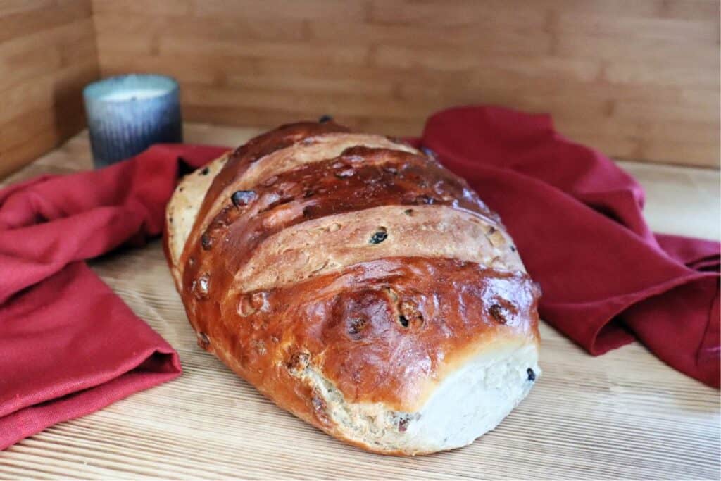 A loaf of apple raisin bread sits on a board. Behind the loaf a red cloth is draped and a candle in a metal holder can be seen. 