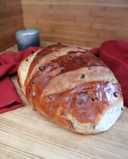 A loaf of apple raisin bread sits on a board. Behind the loaf a red cloth is draped and a candle in a metal holder can be seen.