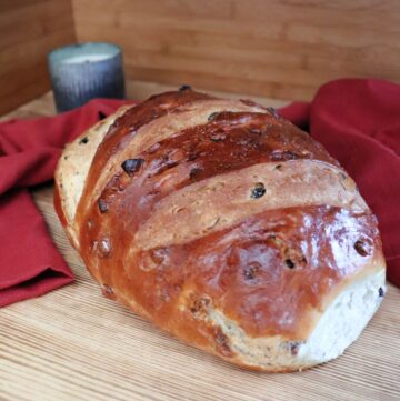 A loaf of apple raisin bread sits on a board. Behind the loaf a red cloth is draped and a candle in a metal holder can be seen.