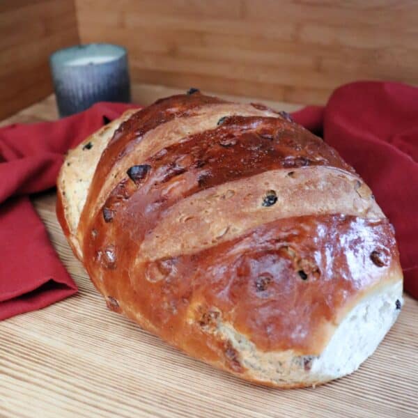 A loaf of apple raisin bread sits on a board. Behind the loaf a red cloth is draped and a candle in a metal holder can be seen.