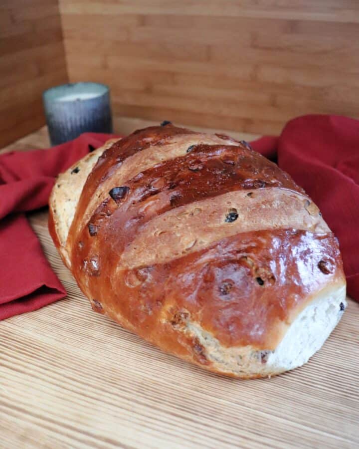 A loaf of apple raisin bread sits on a board. Behind the loaf a red cloth is draped and a candle in a metal holder can be seen.