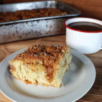 A slice of streusel topped coffee cake sits on white plate with a coffee cup and remaining cake in a pan behind it.