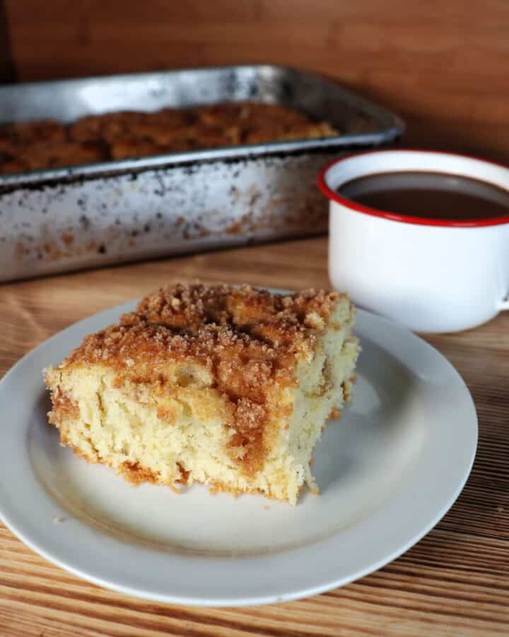 A slice of streusel topped coffee cake sits on white plate with a coffee cup and remaining cake in a pan behind it.