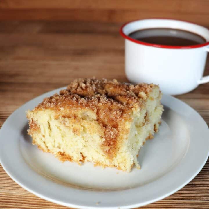 A slice of coffee cake with cinnamon streusel sits ona plate, a white enamel coffee cup full of coffee sits behind it.