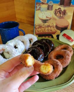 A woman's hand holds half a potato donut covered in cinnamon sugar. Behind it is a platter with a variety of donuts on it.