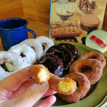 A woman's hand holds half a potato donut covered in cinnamon sugar. Behind it is a platter with a variety of donuts on it.