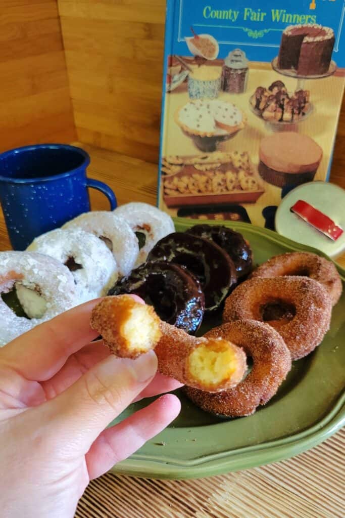 A woman's hand holds half a potato donut covered in cinnamon sugar. Behind it is a platter with a variety of donuts on it. 
