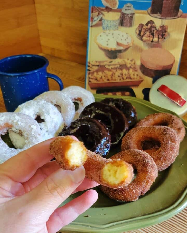 A woman's hand holds half a potato donut covered in cinnamon sugar. Behind it is a platter with a variety of donuts on it.