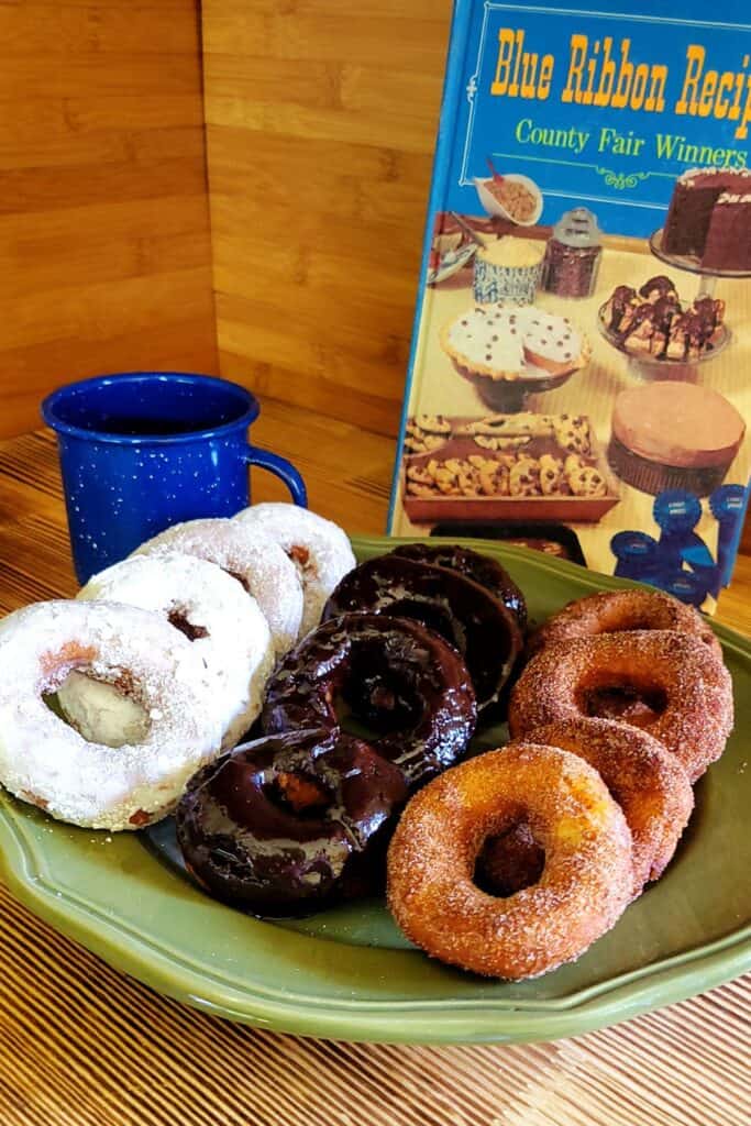 Donuts covered in a variety of toppings sit on a green platter. 
Behind it is a blue tin coffee cup and a vintage cookbook.