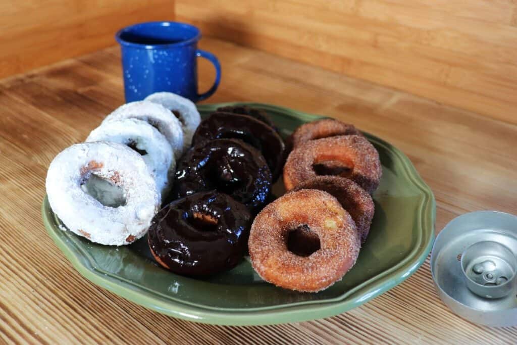 Donuts covered in a variety of toppings sit on a green platter. Next to the platter is an old-fashioned donut cutter and behind it is a blue tin coffee cup. 