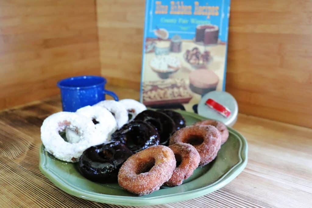 Donuts covered in a variety of toppings sit on a green platter. Behind it is a blue tin coffee cup, a vintage donut cutter, and a vintage cookbook. 